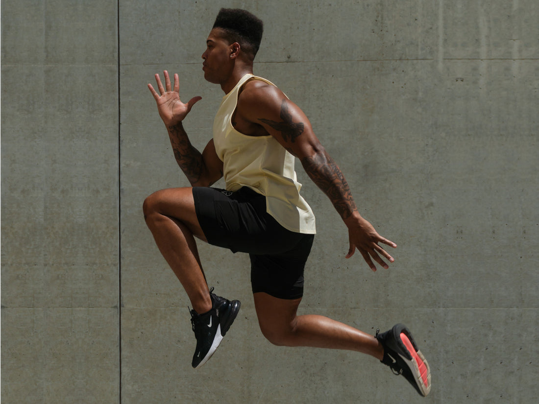 Black man with flat top leaps in the air wearing black running shorts and yellow tank top