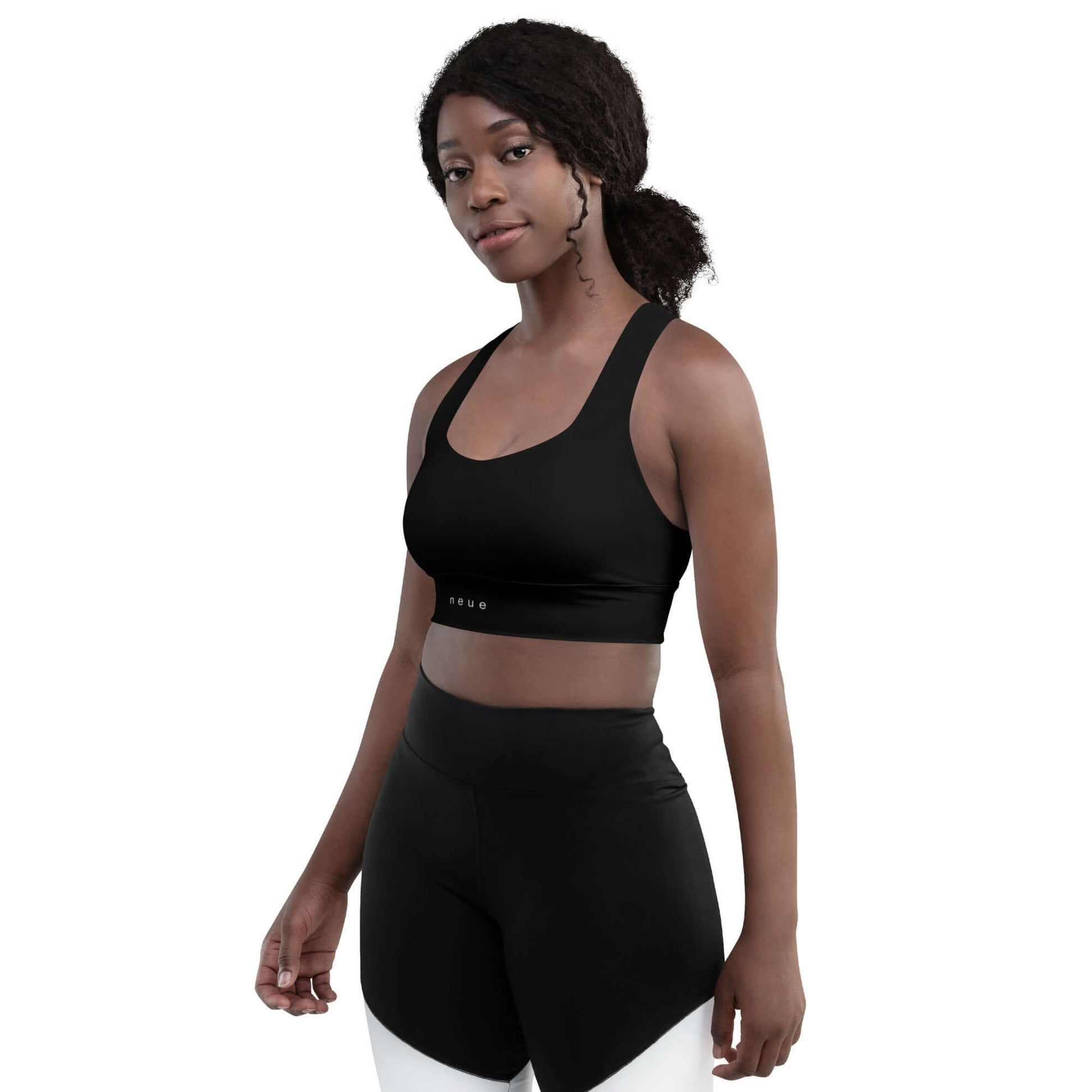 Woman wears Neue Supply Co. Longline Sports Bra in Black. 3/4 view of model looking into camera with white background.