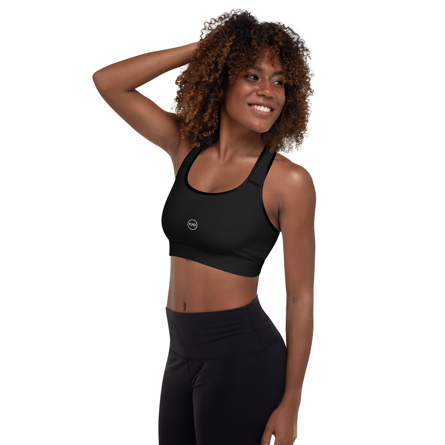 Woman wears Neue Supply Co. Essential Padded Sports Bra in Black. 3/4 view of model smiling with hand on back of head.